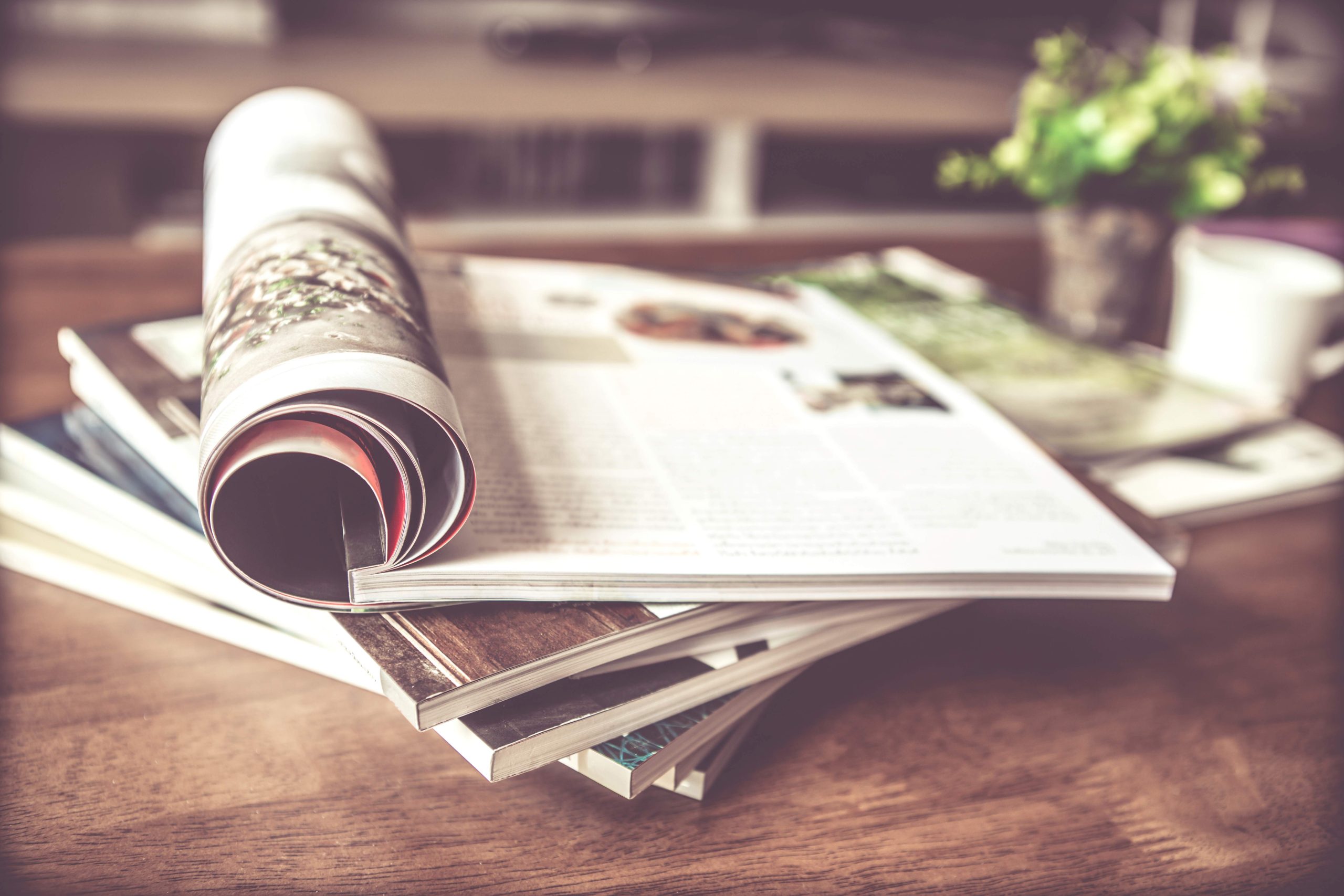 A pile of magazines on a desk. The top magazine is open and is folder over so a reader could be searching for an article.