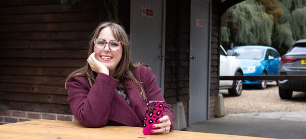 Amy Dawson of Gatekeeper Communications in Ipswich, freelance PR professional sitting at a wooden picnic table outdoors holding a pink phone with black spots