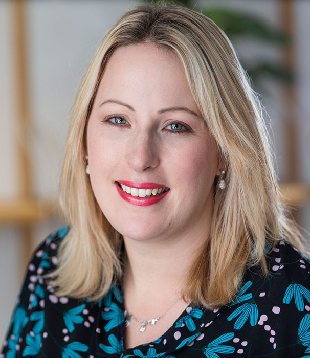 Amy Dawson from Gatekeeper Communications with blonde hair wearing a dark top with teal leaf and pink dot pattern, accessorized with a silver necklace and earrings, in an indoor setting with a blurred background.
