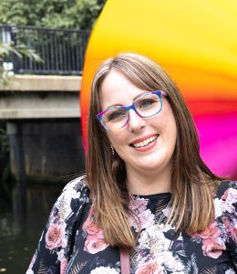 "Amy Dawson of Gatekeeper Communications in Ipswich, freelance PR specialist holding a vibrant rainbow umbrella near a riverside bridge."