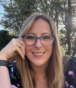 Amy Dawson, a freelance PR and copywriter from Ipswich based Gatekeeper Communications is outdoors wearing a floral-patterned top, with long straight hair, sitting in natural sunlight with trees in the background.