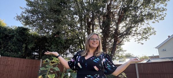 Amy Dawson a report writer from Gatekeeper Communications standing outdoors in a garden, posing with arms outstretched in front of a large tree.
