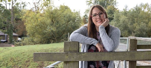 Amy Dawson a freelance PR and copywriter in Ipswich wearing a grey cardigan leaning on a wooden railing near a grassy area with trees in the background."