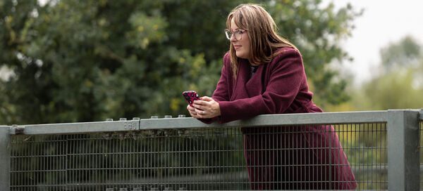 Amy Dawson, an Ipswich copywriter wearing a burgundy coat leaning on a metal railing outdoors while holding a smartphone, with green trees in the background.