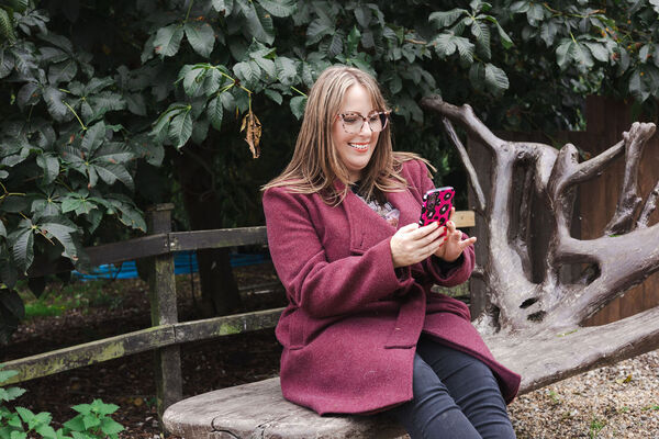Amy Dawson, a freelance PR and copywriter in Ipswich wearing a burgundy coat sitting on a rustic wooden bench outdoors, holding a pink phone with black spots.