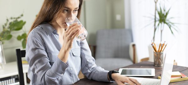 Person in a striped shirt sitting at a desk with a laptop, drinking water in a home office setting