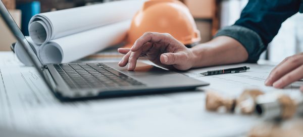 Person working on a laptop with architectural plans and a safety helmet on a desk