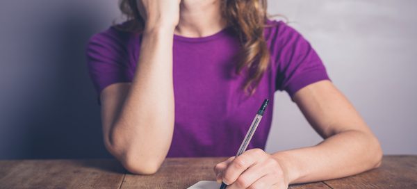 Person in a purple shirt sitting at a wooden table, holding a pen and writing in an open notebook."